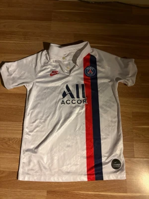 PSG vit matchtröja från Nike - Säljer en vit PSG matchtröja från Nike med korta ärmar och krage. Tröjan har röda och marinblå ränder på framsidan, PSG-logga på bröstet och Nike-logga i rött. Tillverkad i polyester, perfekt för fotboll eller träning.