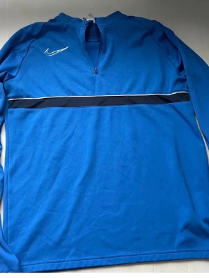Blå långärmad träningströja Nike - Blå långärmad träningströja från Nike med halv dragkedja framtill och vit logga på bröstet. Tröjan har en svart och vit rand tvärs över bröstet och är tillverkad i ett mjukt funktionsmaterial. Perfekt för träning eller uppvärmning.