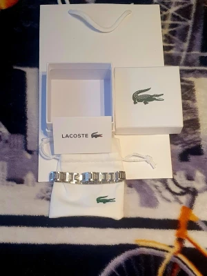 Silverfärgat armband från Lacoste - Snyggt armband i silverfärgad metall från Lacoste med länkdesign och detaljerade länkar. Armbandet har den klassiska Lacoste-loggan och kommer med original ask, påse och presentpåse. Perfekt accessoar för dig som gillar stilrena smycken med sportig touch.
