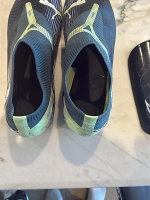 Nike Mercurial fotbollsskor + benskydd - Säljer ett par puma future fotbollsskor som är gröna och svarta med blåa linjer. Har tyvärr inga snören men går att lägga till. Plus jag säljer ett par Nike benskydd som är svarta och har en liten stil av vit.