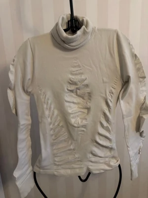 SiSi Vit polotröja med detaljer M - Trendig vit polotröja med unika rynkade detaljer framtill, baktill och längs ärmarna. Tröjan har lång ärm och en hög polokrage i ribbad kvalitet. Perfekt för dig som vill sticka ut med en edgy och modern stil.