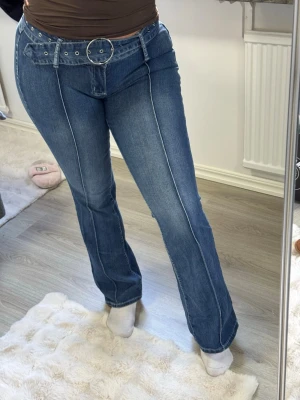 Blå bootcut jeans med bälte - Säljer ett par blå bootcut jeans med markerad söm framtill och tillhörande bälte med rund metallspänne. Jeansen har hög midja och klassisk femficksdesign. Perfekta för en trendig och avslappnad look.