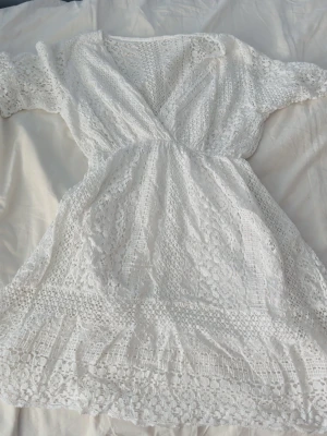 Vit spetsklänning med v-ringning - Säljer en vit kortklänning i spets med v-ringad hals och korta ärmar. Klänningen har omlottkänsla upptill och fina spetsdetaljer över hela plagget. Perfekt för sommarens alla tillfällen!