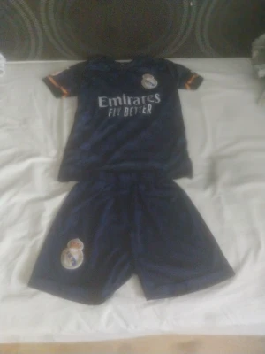 Real Madrid mörkblått matchställ - Snyggt mörkblått matchställ från Real Madrid med klubbmärke på både tröja och shorts. Tröjan har korta ärmar med orange detaljer och trycket 'Emirates Fly Better' på bröstet. Perfekt för fotbollsträning eller att representera favoritlaget. 
