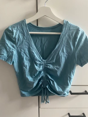 Blå croppad topp med snörning - Ljusblå croppad topp med v-ringning och snörning framtill som ger en snygg rynkad effekt. Kortärmad och tillverkad i mjuk bomull, perfekt för en avslappnad och cool stil. Storlek S men skulle säga XS