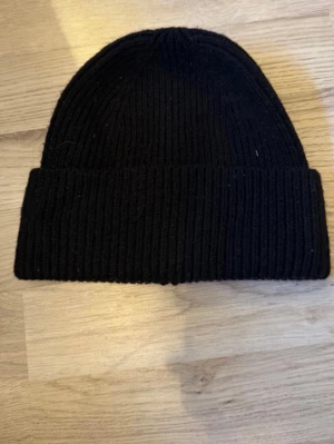 Svart ribbstickad mössa - Säljer en klassisk svart ribbstickad mössa med uppvikt kant. Perfekt för kalla dagar och enkel att matcha med olika outfits. Mössan har en tidlös design och är gjord i mjukt material som sitter skönt på huvudet.