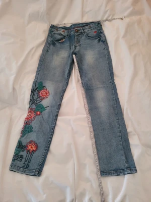 Blå jeans med broderade blommor - Snygga blå jeans med raka ben och broderade röda och gröna blommor längs vänster ben. Jeansen har klassisk femficksmodell, slitningar och kontrastsömmar. Perfekta för dig som vill sticka ut med coola detaljer och unik stil. Saknar 2 knappar. Men den klurige fixar enkelt. 