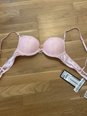 Ljusrosa push-up bh från Censored - Söt ljusrosa push-up bh från Censored med små broderade rosetter på kuporna och dekorativ rosett i mitten. Bh:n har justerbara axelband och detaljer i satinliknande material. Perfekt för dig som gillar feminina detaljer.