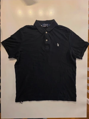 Svart pikétröja från U.S. Polo Assn. - Klassisk svart pikétröja från U.S. Polo Assn. med broderad logga på bröstet. Tröjan har krage, korta ärmar och knäppning med tre knappar framtill. Perfekt för en clean och sportig stil. Väldigt snygg och sportig vid biceps          STORLEK M
