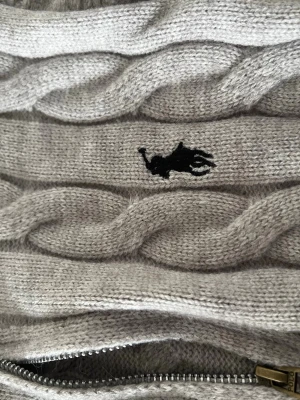 Grå kabelstickad tröja Polo Ralph Lauren - Snygg grå kabelstickad tröja från Polo Ralph Lauren med halv dragkedja och svart broderad logga på bröstet. Tröjan har ribbade muddar och hög krage, perfekt för kyliga dagar. Klassisk och tidlös design i mjukt material.