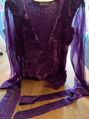 Lila omlottopp från Serena Kay - Unik lila omlottopp från Serena Kay med transparenta långärmar och dekorativa spetsdetaljer framtill. Toppen har v-ringning och långa band för knyt i midjan. Perfekt för dig som vill sticka ut med en kreativ och konstnärlig look.