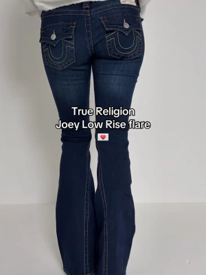 True Religion Joey Low Rise flare jeans - Mörkblå jeans från True Religion, modell Joey Low Rise flare. Jeansen har låg midja, utsvängda ben och dekorativa fickor bak med kontrastsömmar och silverfärgade knappar. Klassisk femficksmodell i denim med snygga detaljer.