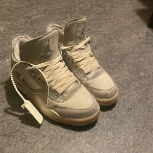 Off-White x Air Jordan 4 Sail sneakers - Säljer ett par Off-White x Air Jordan 4 Sail sneakers i beige och grå toner. Skorna har meshpaneler, synliga sömmar, Off-White tag och klassisk Jumpman-logga på plösen. Stilren design med högt skaft och robust sula. Perfekta för dig som gillar streetwear och unika detaljer.