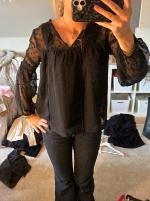 Svart transparent blus med prickar - Snygg svart blus i transparent material med diskreta prickar och spetsdetaljer vid halsringningen. Blusen har långa, luftiga ärmar och en lös passform som ger en avslappnad men trendig look. Perfekt att styla med jeans eller kjol.