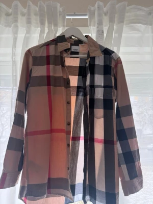Rutig skjorta från Burberry - Säljer en klassisk rutig skjorta från Burberry i beige, svart, vit och röd. Skjortan har långa ärmar, knappar framtill och en bröstficka. Tillverkad i tunn bomull som känns skön mot huden. Perfekt för dig som gillar tidlös och ikonisk stil.