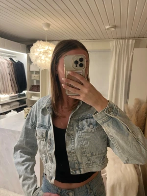 Cropped ljusblå jeansjacka - Trendig cropped jeansjacka i ljusblå denim med klassisk krage, bröstfickor med lock och knappstängning framtill. Jackan har en avslappnad passform och långa ärmar med knapp vid ärmslut. Perfekt att styla med höga jeans eller kjol.