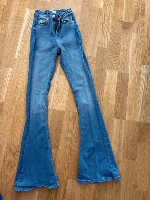 Blå utsvängda jeans från Gina  - Säljer ett par blå jeans från gina med hög midja och utsvängda ben. Klassisk femficksmodell med bälteshällor och dragkedja med knapp. Jeansen är i mjukt denimtyg och har en snygg tvättad look. Perfekta för dig som gillar retrostil och vill sticka ut. Storlek xs