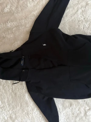 Mörkblå hoodie från Polo Ralph Lauren - Klassisk mörkblå hoodie från Polo Ralph Lauren med vit broderad logga på bröstet. Hoodien har dragsko i huvan och en stor magficka framtill. Tillverkad i mjukt bomullsmaterial och har en avslappnad passform.