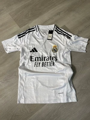 Real Madrid matchtröja Adidas vit - Säljer en vit Real Madrid matchtröja från Adidas med svarta detaljer och klubbmärke på bröstet. Tröjan har svarta ränder på axlarna och trycket 'Emirates Fly Better' på framsidan. Perfekt för fotbollsfans som vill bära sitt lag med stil.