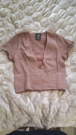 Ribbad croppad topp Urban Outfitters - Säljer en ribbad croppad topp från Urban Outfitters i en varm brun nyans. Toppen har djup v-ringning och korta ärmar, perfekt för en trendig och avslappnad stil. Materialet är mjukt och stretchigt för extra komfort.