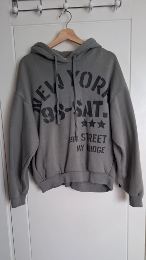 Grå, grön aktig hoodie från Bershka  - Snygg grå hoodie med tryck på både fram- och baksidan. Texten inkluderar 'NEW YORK', 'BROOKLYN' och andra coola detaljer. Hoodien har huva med dragsko och breda muddar vid ärmslut och nederkant. Perfekt för en avslappnad streetstyle-look.