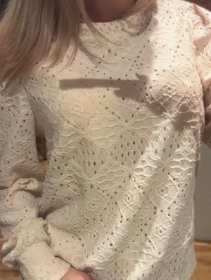 Krämvit spetsblus med lång ärm - Superfin krämvit blus i spets med broderat mönster och lång ärm. Blusen har rund halsringning och är tillverkad i ett lätt, luftigt material som ger en romantisk vibe. Perfekt att styla med jeans eller kjol för en trendig look.