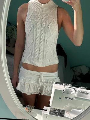 Vit stickad croppad tröja - Snygg vit stickad väst med grovt kabelstickat mönster och ribbad hög krage. Modellen är ärmlös och croppad, perfekt att styla över en skjorta eller t-shirt. Materialet är mjukt och ger en trendig look. 