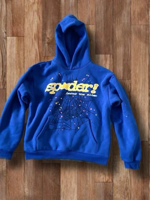 Blå Spi5der hoodie  - Blå hoodie med huva och trycket 'spider! break the rules' i gult framtill. Stort spindelnätsmotiv och små färgglada stjärnor på både fram- och baksida. Mjuk bomullskänsla och ribbade muddar vid ärmar och midja. Perfekt för dig som gillar streetwear och coola prints.