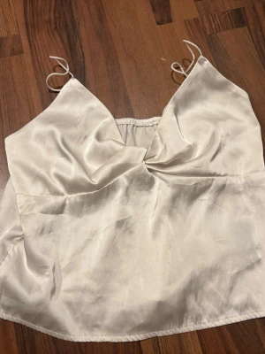Vit satin linnetopp - Snygg linnetopp i satin med tunna axelband och lätt draperad front. Toppen har en croppad passform och ger en lyxig känsla med sitt glansiga material. Perfekt att styla med jeans eller kjol för en trendig look. Storlekslapp bortklippt men är storlek S. Använd en gång. 