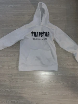 Vit hoodie från Trapstar - Säljer en vit hoodie från Trapstar med svart text 'TRAPSTAR IT'S A SECRET' på bröstet. Hoodien har en stor magficka och klassisk huva. Tillverkad i mjukt material, perfekt för en avslappnad streetwear-look.