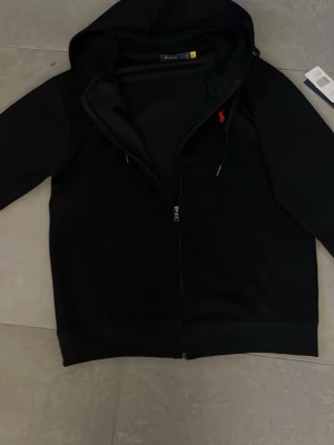 Svart hoodie från Polo Ralph Lauren - Svart hoodie med dragkedja från Polo Ralph Lauren. Klassisk design med huva och dragsko, samt röd broderad logga på bröstet. Tillverkad i mjukt material och har fickor framtill. Perfekt för en avslappnad och stilren look. Köpt ny men passar tyvär inte mig!