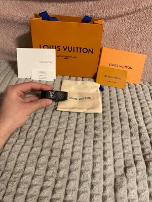 Louis Vuitton svart läderarmband - Snyggt svart armband från Louis Vuitton i äkta läder med klassiskt LV-monogram i silverfärgad metall. Armbandet har en stilren design och levereras med originalpåse och förpackning. Perfekt accessoar för att lyfta din outfit.