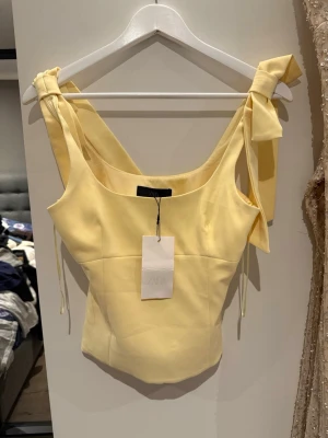 Gul linnetopp med rosetter från Zara - Supersöt gul linnetopp från Zara med breda axelband och stora rosetter på axlarna. Toppen har en figurnära passform och är perfekt för varma dagar. Materialet känns mjukt och lätt, och färgen är riktigt somrig.