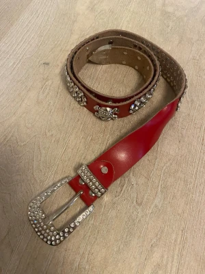 Rött dödskalle skärp  - Coolt rött bälte med massor av glittrande strass och nitar längs hela bandet. Spännet är dekorerat med stenar för extra bling. Vet tyvärr inte märke, är köpt på plick💞💞105cm långt!