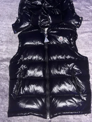 Svart dunväst från Moncler - Snygg svart dunväst från Moncler med glansig finish och ikonisk logga på bröstet. Västen har dragkedja framtill, två fickor med dragkedja och hög krage. Insidan har klassisk Moncler-serietidning och etikett. Perfekt för lager-på-lager och streetstyle.