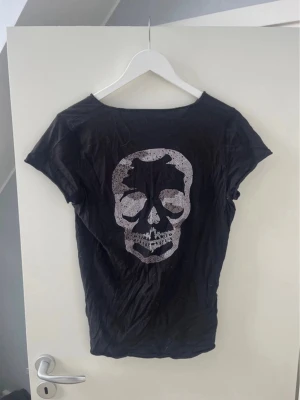 Svart t-shirt med döskalletryck - Cool svart t-shirt med ett stort döskalletryck i silver på ryggen och v-ringning framtill med små knappar längs kanten. T-shirten har korta ärmar och är gjord i mjukt material, perfekt för en edgy och avslappnad stil.