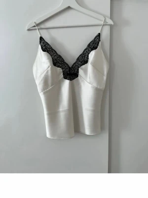 Vit satinlinnetopp med spetsdetalj - Snygg vit linnetopp i satin med smala axelband och svart spets längs v-ringningen. Toppen har en figurnära passform och ger en lyxig känsla med sitt blanka material och eleganta spetskant. Perfekt att styla med jeans eller kjol för en trendig look.