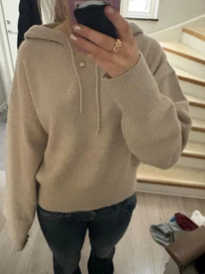 Beige hoodie i mjuk stickad kvalitet - Säljer en beige hoodie med huva och dragsko. Tröjan är stickad i ett mjukt material och har ribbade muddar vid ärmslut och nederkant. Perfekt för en avslappnad och stilren look. Passar till jeans eller mjukisbyxor. I storlek 170