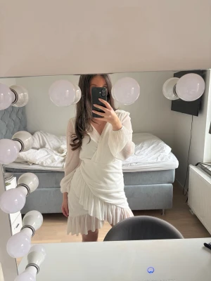 Vit omlottklänning från Vila - Supersnygg vit total omlottklänning från Vila med långärmade, lätt transparenta ärmar och volangdetaljer vid kjolen segmentet. Klänningen har v-ringning och kort, figurnära passform som ger en elegant vibe. Materialet är mjukt och faller mailt. Perfekt för dig som vill sticka ut.