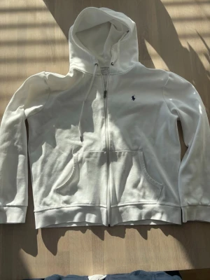 Vit hoodie från Polo Ralph Lauren - Snygg vit hoodie med dragkedja från Polo Ralph Lauren. Klassisk design med huva och dragsnören, två fickor framtill och den ikoniska blå loggan broderad på bröstet. Tillverkad i mjukt bomullsmaterial som är skönt att ha på sig.
