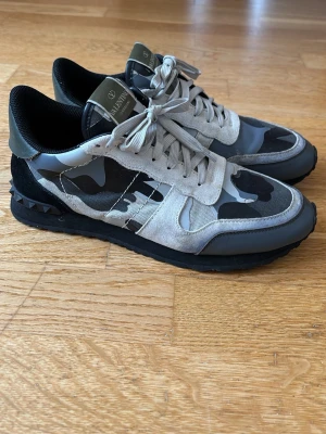 Valentino camo sneakers i grått - (Pris kan diskuteras)! Snygga sneakers från Valentino med grå och svart camo-mönster, mockadetaljer och svarta nitar på hälen. Skorna har en låg silhuett, vita och grå skosnören samt svart yttersula. Perfekta för dig som vill sticka ut med fina skor, lite använda och lite småskromor därför sänkt pris. 