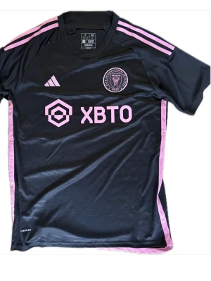 Inter Miami matchtröja svart/rosa Adidas 23/24 - Snygg svart matchtröja från Inter Miami CF med rosa detaljer och Adidas-logga. Tröjan har klubbmärke på bröstet, sponsortryck och tre rosa ränder på axlarna. Tillverkad i ett lätt och ventilerande funktionsmaterial, perfekt för träning eller match.