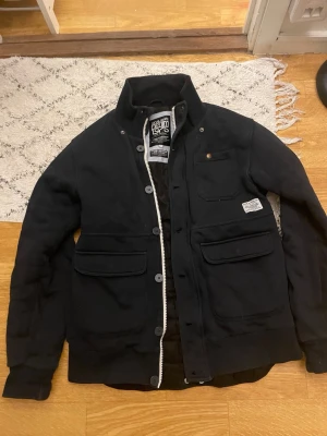 Svart jacka från Denim & Supply Ralph Lauren - Stilren svart jacka från Denim & Supply Ralph Lauren med hög krage, flera fickor framtill och knapp- samt dragkedjestängning. Jackan har patchdetaljer och är tillverkad i ett kraftigare bomullsmaterial. Perfekt för dig som gillar en klassisk men cool look.