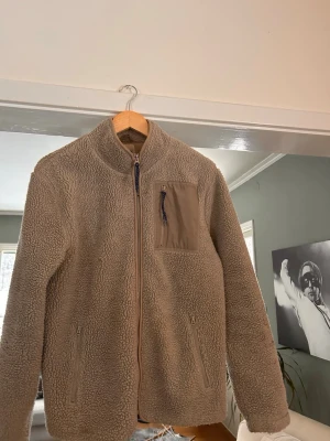 Beige teddyjacka med bröstficka - Skön jacka som man enkelt kan ha oavsett årstid. Våldigt bra skick men en liten fläck på ärmen som inte syns. Passar perfekt på 170-180.