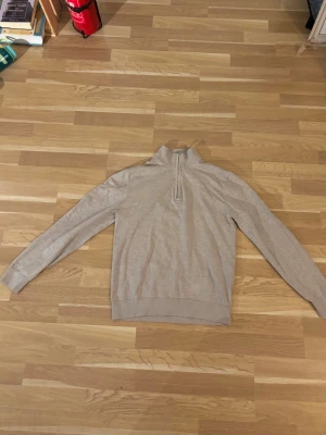 Beige halvzip-tröja från Bläck - Snygg beige tröja från Bläck med halv dragkedja och hög krage. Tillverkad i mjukt material och har ribbade muddar vid ärmslut och nederkant. Perfekt för en stilren och avslappnad look.