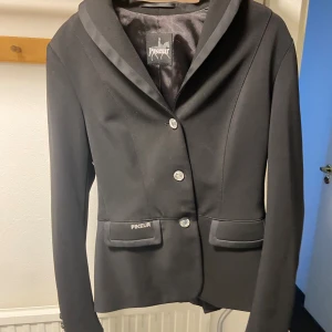 Svart ridkavaj från Pikeur - Stilren svart kavaj från Pikeur med klassisk krage och tre silverfärgade knappar framtill. Kavajen har två fickor med lock och Pikeur-logga på ena fickan. Snygg, figursydd passform och slätt tyg som ger en elegant look.