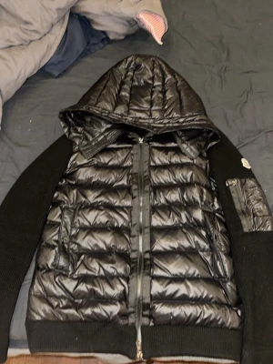 Svart cardigan jacka från Moncler - Svart cardigan jacka från Moncler med quiltad framsida i dun och stickade ärmar. Jackan har huva med snörning, dragkedja framtill och Moncler-logga på ärmen. Perfekt för dig som vill ha en stilren och sportig look.