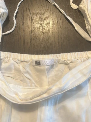 Vit offshoulder blus från Flash - Söt vit offshoulder blus från Flash med puffiga ärmar och tunna knytband vid axlarna. Blusen har resår upptill och fina veckdetaljer på ärmarna. Tillverkad i ett lätt och luftigt tyg, perfekt för en trendig och avslappnad look.