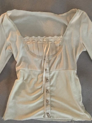 Ljusbeige ribbad topp med spetsdetalj - Benvit Topp som jag köpte för några år sedan som jag säljer nu pga den kommer ej till användning längre! Den har lång ärm och ett band att knyta framtill för extra detalj. En liten defekt nertill se bild 2)