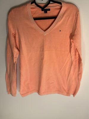 Persikofärgad tröja Tommy Hilfiger - Persikofärgad långärmad tröja från Tommy Hilfiger med v-ringning och liten broderad logga på bröstet. Tröjan är i mjukt material och har ribbade muddar vid ärmslut och nederkant. Perfekt för en stilren och avslappnad look.